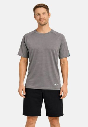 Mann trägt ein graues YETI-T-Shirt mit kurzen Ärmeln und schwarze Shorts, steht mit entspannter Haltung vor einem schlichten Hintergrund.