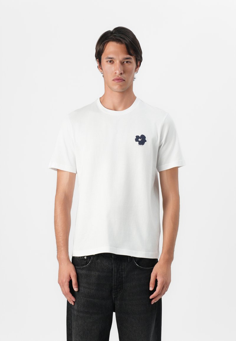 White cotton t-shirt with a round neck and short sleeves. Features a small navy graphic on the left chest. Paired with black trousers.