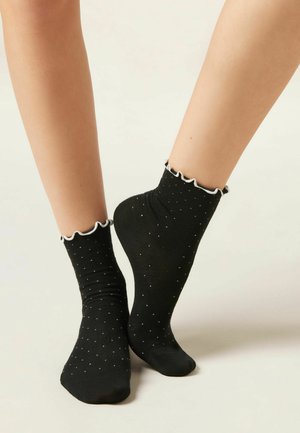 Gambe con calzini alla caviglia neri con piccoli puntini bianchi e rifinitura arricciata bianca, posizionate su uno sfondo neutro chiaro.