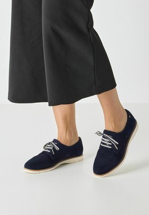 Marineblauwe suède schoenen met witte veters, geperforeerd ontwerp, rubberen zool en een tankleurig detail bij de neus, gecombineerd met zwarte, loszittende broek.