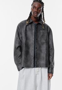 Sort læderagtig knap-op jakke med en frontlomme, afslappet pasform og kontrasterende syninger. Bæres over grå sweatpants.