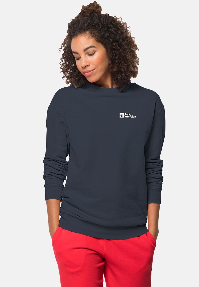 Felpa girocollo blu navy realizzata in tessuto morbido, con maniche lunghe e un piccolo logo bianco sul petto. Abbinata a pantaloni rossi.