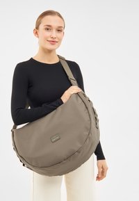 Borsa a tracolla verde oliva di forma rotonda, realizzata in tessuto morbido. Presenta una tracolla intrecciata e un'etichetta cucita con il logo sul davanti.