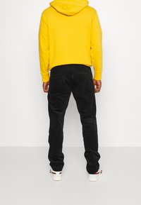 Pantaloni in corduroy neri con vestibilità slim e due tasche posteriori, abbinati a una felpa con cappuccio gialla. Sneakers bianche con dettagli colorati.