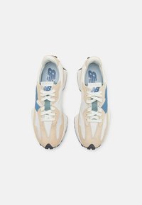 Paire de baskets New Balance beige, blanches et bleues avec des lacets blancs, vues de dessus sur un fond blanc uni.