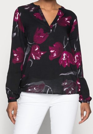 Sort langærmet bluse med lilla blomsterprint, båret med hvide bukser, der viser en V-hals og opsamlede manchetter på ærmerne.