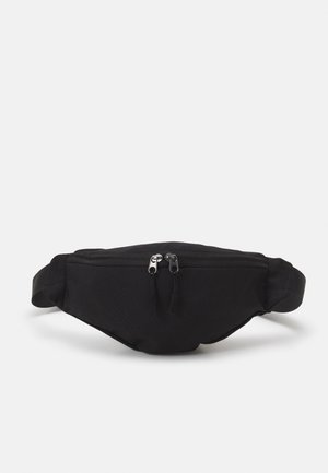 Sac banane en tissu noir avec une poche zippée à l'avant comportant deux tirettes de fermeture éclair argentées et une sangle réglable sur fond blanc.