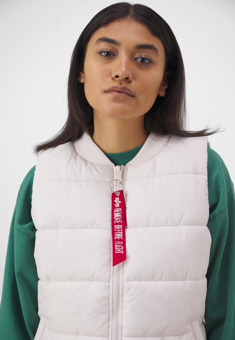 Helles pink gepolstertes Westchen mit einem Reißverschluss, geripptem Kragen und einem roten Stoffetikett, auf dem steht "VOR DEM FLUG ENTFERNEN". Getragen über einem grünen Oberteil.