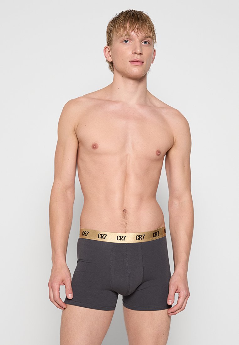 Junger Mann mit kurzen blonden Haaren, der dunkelgraue Boxershorts mit goldenem CR7-Bund trägt und vor einem hellen, einfarbigen Hintergrund steht.