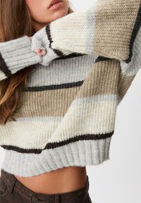 Pletenji pulover s horizontalnim prugama u bež, sivoj i bijeloj boji. Rebrasti rukavi i rub s laganim skraćenjem, koji naglašava mekanu teksturu.
