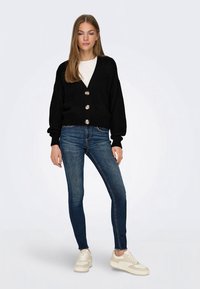 Cardigã de malha preta com três botões grandes, combinado com jeans azuis de corte justo e ténis brancos. Textura suave, ajuste descontraído, design canelado.
