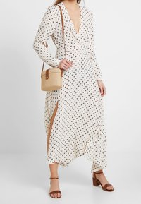 Lång, krämfärgad klänning med bruna prickar, djup V-ringning, långa ärmar, sidslitsar, i kombination med en vävd väska och bruna klacksandaler.