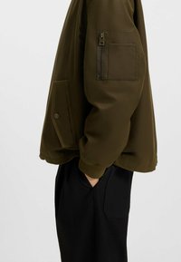 Olive green bomber jacket with a zippered sleeve pocket and side patch pocket. Ribbed cuffs and hem, made of smooth, lightweight fabric.