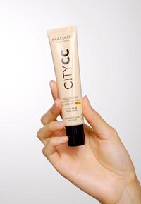 Un tubetto beige di CITYCC Hyaluronic Anti-Pollution CC Cream SPF 15, con un tappo nero e un design elegante, tenuto in mano su uno sfondo chiaro.