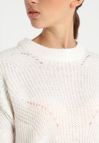 Maglione bianco a maglia con scollatura a girocollo, motivo a maglia aperta con piccoli fori e una finitura strutturata.