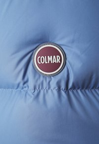 Šviesiai mėlynas pūkuotas striukės audinys su apvaliu bordo ir baltu Colmar logotipu fronto dalyje, pasižymintis lygiu tekstūru ir siūlėmis detalėmis.