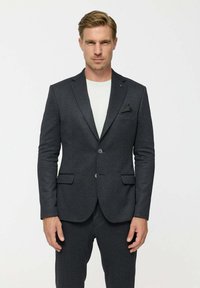 Férfi áll előre nézve, sötétszürke öltönykabáttal és nadrággal, fehér inggel alatta, egyszerű világos háttér előtt.