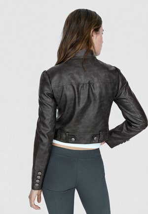 Frau mit langen Haaren, von hinten gesehen, trägt eine eng anliegende dunkelbraune Lederjacke und dunkle Leggings vor einem einfarbigen hellen Hintergrund.