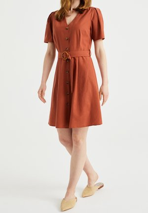 Frau trägt ein rostfarbenes Kleid mit Knopfleiste, passendem Gürtel und kurzen Ärmeln, kombiniert mit beigen, geflochtenen Slip-on-Schuhen.