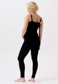 Blonde vrouw die glimlacht, staand op blote voeten, iets zijwaarts gedraaid, in een zwart tanktop en legging tegen een neutrale witte achtergrond.