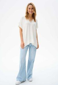 Witte V-hals top met kantafwerking, relaxte pasvorm, losse mouwen, gecombineerd met lichtblauwe denim flared broeken en witte sneakers, tegen een neutrale achtergrond.