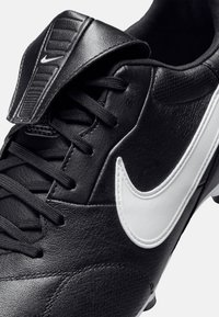 Chaussure de football en cuir noir avec des lacets noirs, une languette rembourrée et un Swoosh Nike blanc sur le côté. Présente des détails en matériau texturé.