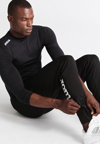 Zwarte sportshirt met lange mouwen van textuurstof en Joma-logo op de schouder; gecombineerd met zwarte broek met een wit Joma-logo.