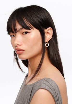 Mujer joven con cabello negro liso y flequillo, vistiendo una camiseta sin mangas gris y pendientes de aro dorados y blancos, mirando de lado.