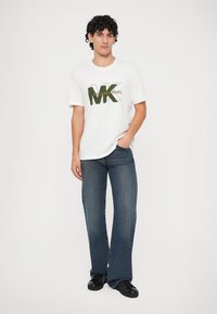 Balta kokvilnas T-kreklā ar zaļu "MICHAEL KORS" logotipu, saskaņots ar tumši ziliem platām biksēm. Modelis valkā melnas kurpes un stāv pret vienkāršu fonu.
