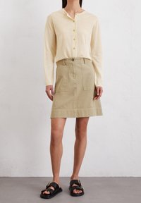 Žena v béžovém knoflíkovém topu s dlouhým rukávem, khaki sukně do kolen s předními kapsami a černých sandálech na přezku, stojící na šedé podlaze.