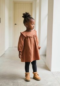 Braun karierte Kleid mit Rüschenausschnitt und langen Ärmeln, schwarze Leggings und hellbraune, gefütterte Stiefel. Neutrale Innenraum-Hintergrund.