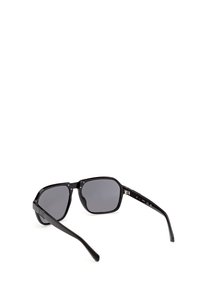 Lunettes de soleil à monture carrée noire avec verres teintés foncés et branches légèrement courbées, reposant ouvertes sur une surface blanche.