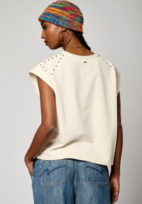 Un haut beige à manches courtes décoré de clous sur les épaules, associé à un jean en denim bleu avec un design de poche cousu.