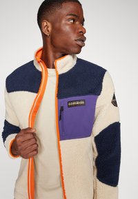 Fleece jacket with cream, navy, and purple sections. Features an orange zip, side pocket, and textured fabric. Elastic cuffs.