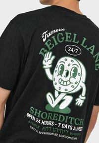 Zwart katoenen T-shirt met een groene en witte cartoon bagel karakter en de tekst: "Famous Beigel Lane 24/7 Shoreditch. 24 uur geopend."