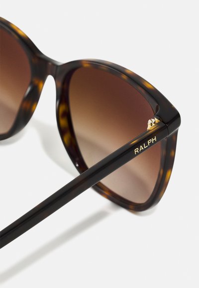 Tortoiseshell sunglasses with gradient brown lenses. Distinctive cat-eye shape, black temple with gold "RALPH" logo and metal accent detail.