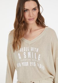 Leicht goldfarbenes V-Ausschnitt-Pullover mit lockerer Passform, langen Ärmeln und weißem Text "REBEL WITH A SMILE" auf der Vorderseite.