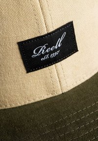 Gorra kaki y verde con tejido texturizado. Presenta una etiqueta negra con texto bordado en blanco que dice "Reell est. 1997." Detalles de costura visibles.