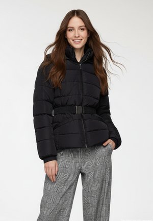 Giacca a vento nera con collo alto, chiusura con zip e vita elasticizzata. Indossata sopra pantaloni ampi a quadri grigi. Texture liscia e design trapuntato.