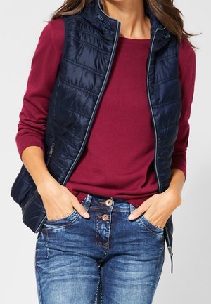 Woman wearing a navy quilted sleeveless vest over a burgundy long-sleeve shirt, with her hands in the pockets of blue jeans.