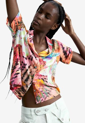 Žena s vrkočmi, oblečená v farebnej kvetinovej krátkoročnej košeli na gombíky a bielych nohaviciach, pózujúca so zatvorenými očami a zdvihnutými rukami.