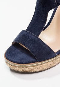 Sandale compensée en daim bleu marine avec une large bride avant et une semelle plateforme en corde tressée sur fond blanc.