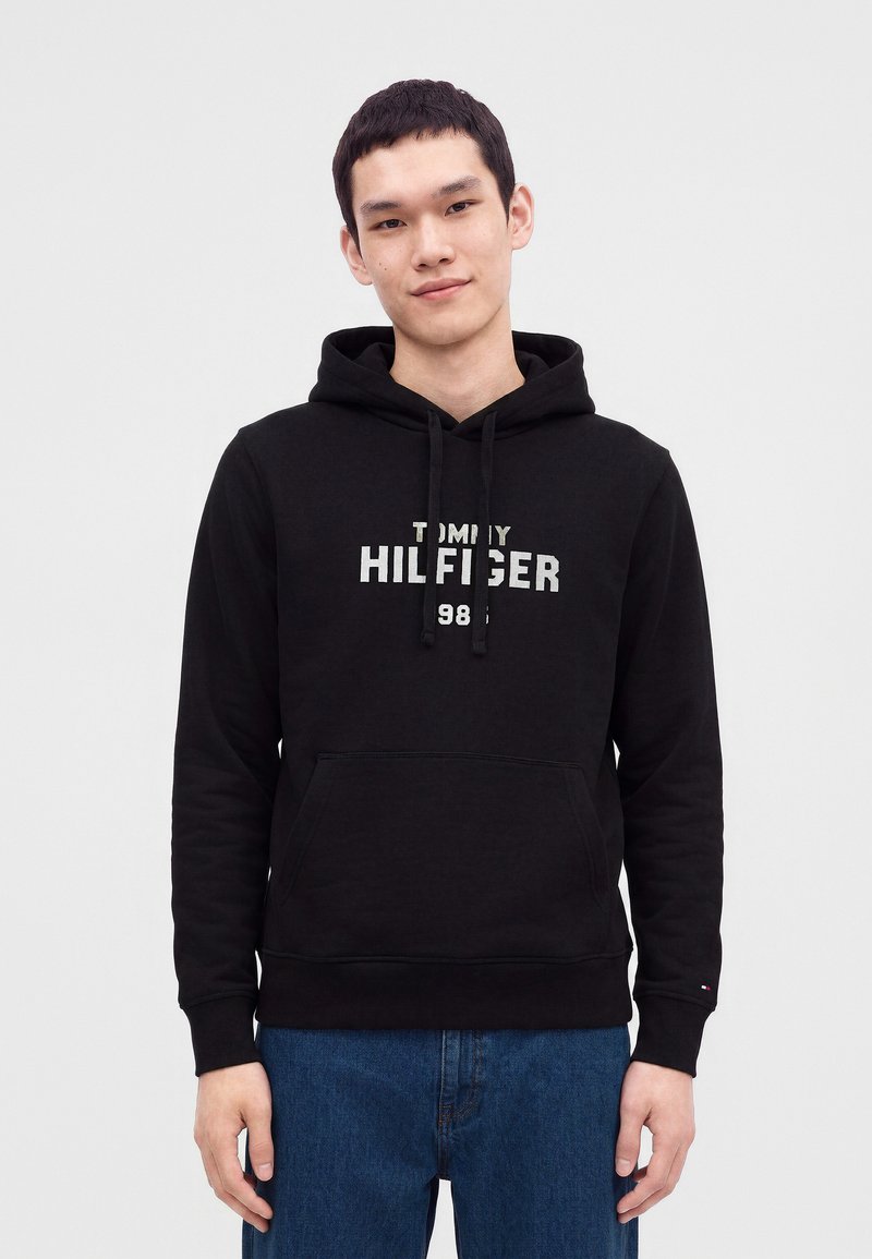 Млад мъж, облечен в черна суитшърт Tommy Hilfiger с лого и син деним, стои пред светлосив фон.