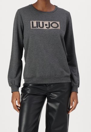 Vrouw draagt een donkergrijze trui met lange mouwen met de tekst "LIU·JO" in strasstenen en zwarte neplederen broek.