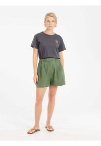 Grijze t-shirt met een kleine grafische afbeelding, gecombineerd met hoge groene shorts. Model draagt tan slippers, geplaatst tegen een witte achtergrond.