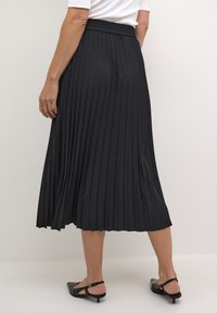 Czarna plisowana spódnica z elastycznym paskiem w talii, o długości do połowy łydki, w płynnym kroju i gładkiej fakturze. Do tego czarne buty na pasku z tyłu.