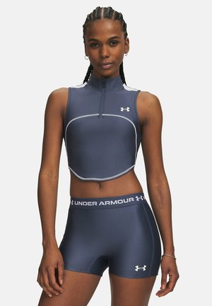 Athletische Frau mit geflochtenem Haar, die ein ärmelloses dunkelblaues Crop-Top und passende Shorts mit Under Armour-Logos trägt.