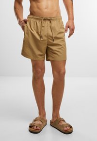 Pantaloni scurți de înot bej, cu talie elastică și șnur. Textură de țesătură netedă. Purtați cu sandale de plajă maro.