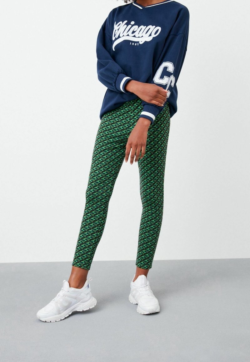 Personne portant un sweat-shirt bleu marine Chicago, un pantalon vert à motif géométrique et des baskets blanches, debout devant un fond uni.
