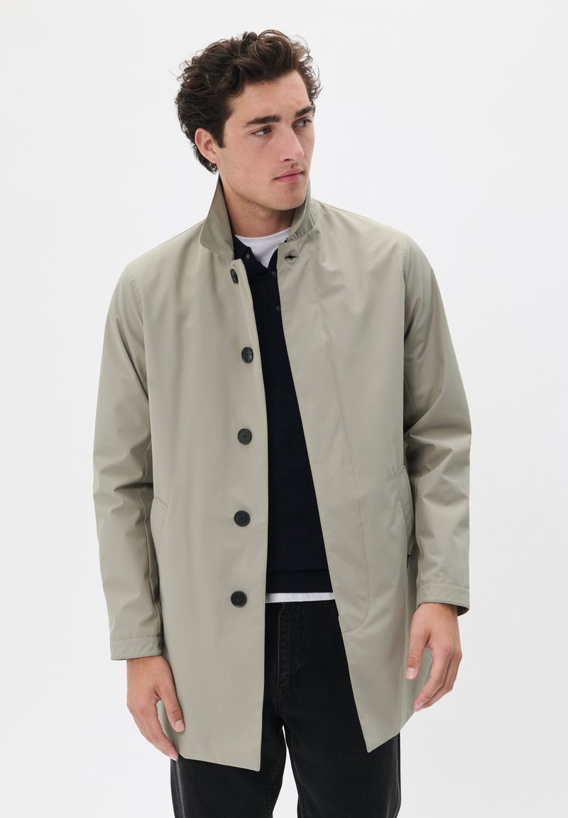 Chaqueta impermeable de color beige claro con cuello, botones negros, bolsillos laterales y un corte relajado. Lleva puesta sobre una camisa azul marino y pantalones negros.